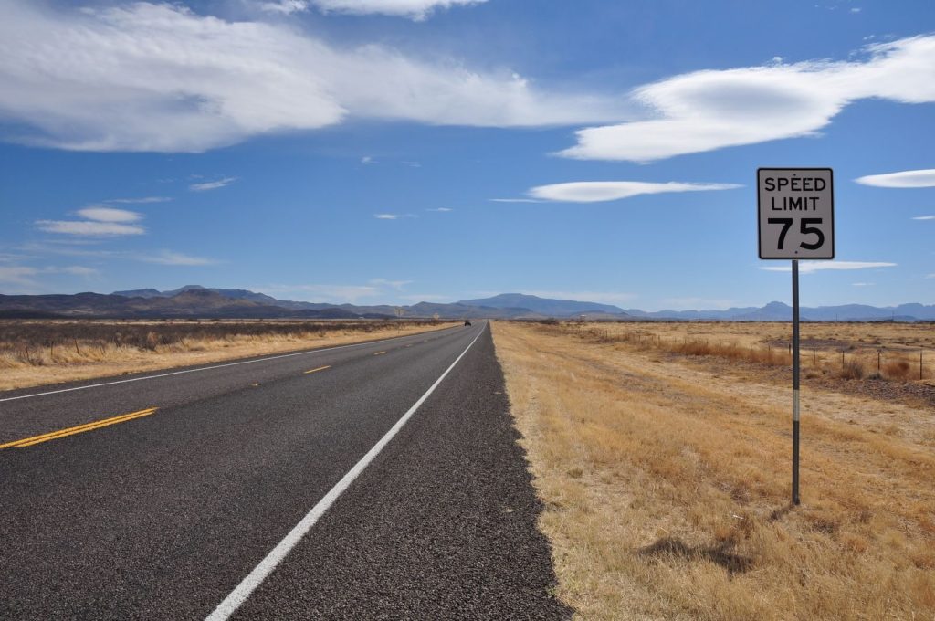A deserted highway stretches into the horizon under a bright blue sky with a speed limit sign in the countryside.