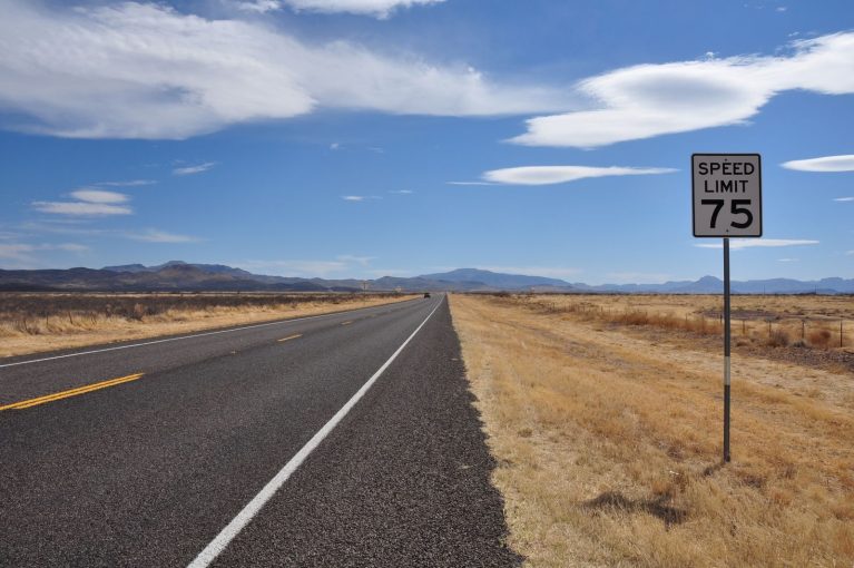 A deserted highway stretches into the horizon under a bright blue sky with a speed limit sign in the countryside.