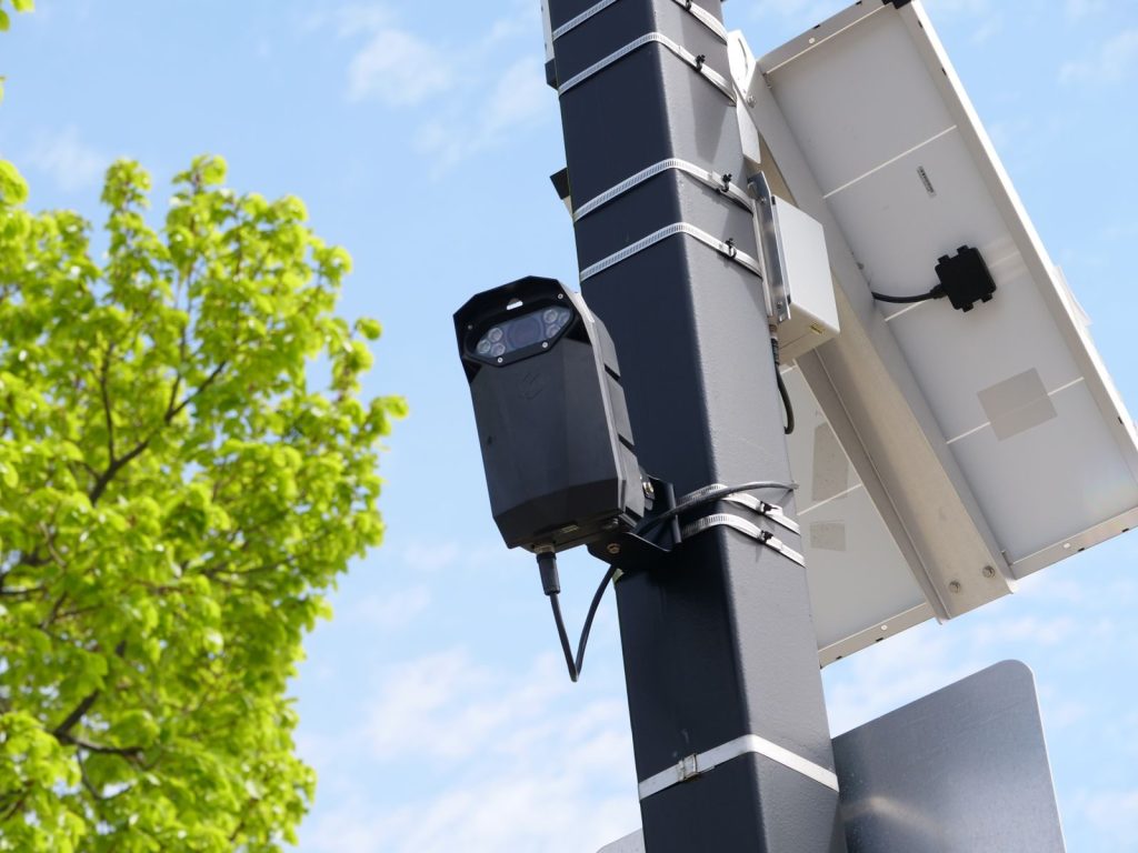 Urban surveillance camera mounted on pole with solar panel and green tree in view.
