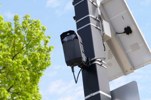 Urban surveillance camera mounted on pole with solar panel and green tree in view.