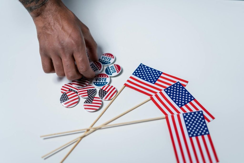A hand reaches for voting buttons and American flags on a white background.