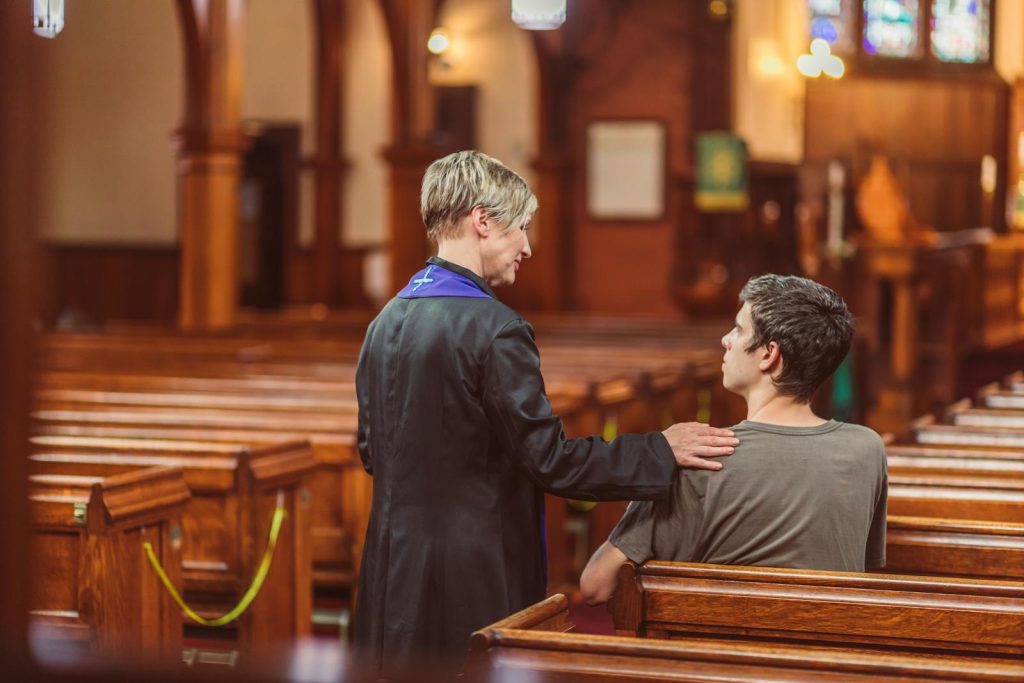 A clergy member offering comfort to a seated individual inside a tranquil church environment.