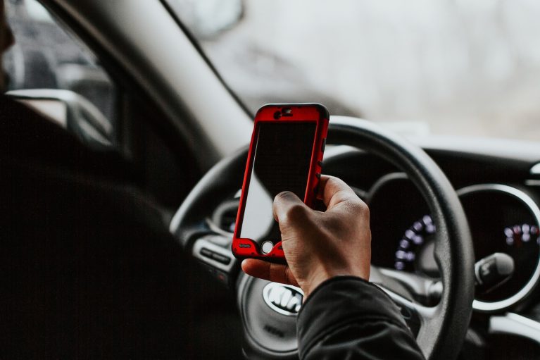 person holding red smartphone sitting in front of vehicle steering wheel